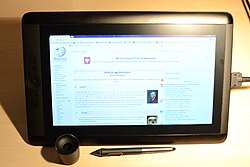 A monitor with the Dutch Wikipedia displayed. A large cable is plugged into the right side of the monitor. The Cintiq is standing upright on a desk, and lower down on the image is a digital pen and the pen holder.