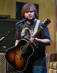 A photograph of Jack Black, in casual clothes, holding an acoustic guitar while on stage.