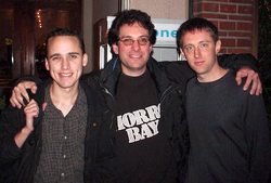 Three dark-haired men wearing dark clothing. The man on the left is wearing a checkered shirt with a rain jacket. The man in the middle is wearing a printed tee with a rain jacket. The man on the right is wearing a black sweater.