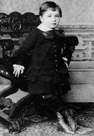 A young boy with short hair and a round face, wearing a white collar and large bow, with vest, coat, skirt, and high boots. He is leaning against an ornate chair.