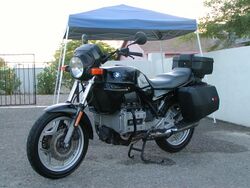 Black BMW K75T with top box and panniers, parked on a driveway in front of a house and metal gates