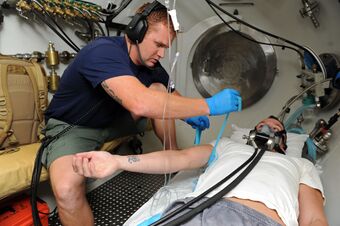 Photograph of the cramped interior of a decompression chamber with a Hospital Corpsman preparing a patient for an intravenous line during a demonstration of patient care for decompression illness.
