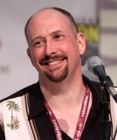A balding, middle aged caucasian male looks slightly to the left with a smile. He is wearing a black and cream shirt with palm tree images upon it.