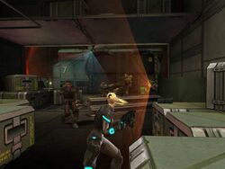 A young blonde woman in an armored suit and carrying a gun runs for cover behind crates while a group of guards take aim, using a sensor device to sweep the area.