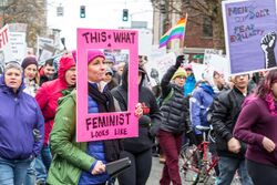 A large group of people in the Seattle Women's March, some holding handwritten signs. The image is focused on one marcher holding a cowbell and a pink cutout sign with the words "this is what a feminist looks like." The sign looks like a picture frame and is outlining the marchers face.