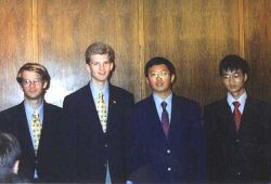 Four men in black suits with bluish-white dress shirts and brightly-coloured ties standing in front of a wall composed of wooden panels.