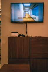 A television screen placed above a placard with a controller next to it. The television is displaying part of the "Confusion Ending" of the remake, in which the player is told to follow a painted yellow line through the office.