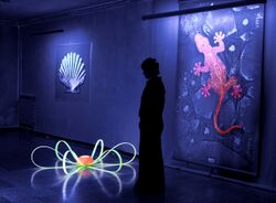 A person in silhouette stands gazing down at a piece of art on the floor. Paintings on the wall behind the person are glowing from the UV light.